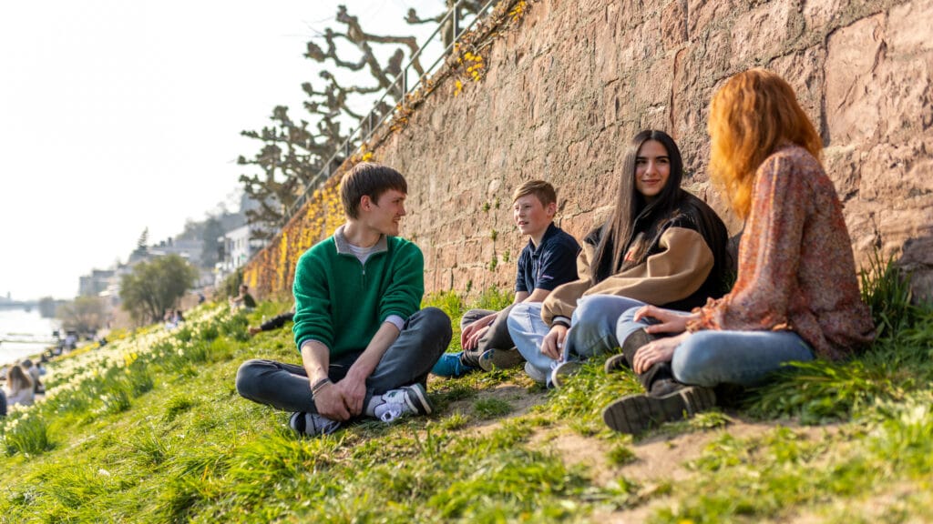 Vier Jugendliche sitzen auf der Neckarwiese und sprechen miteinander