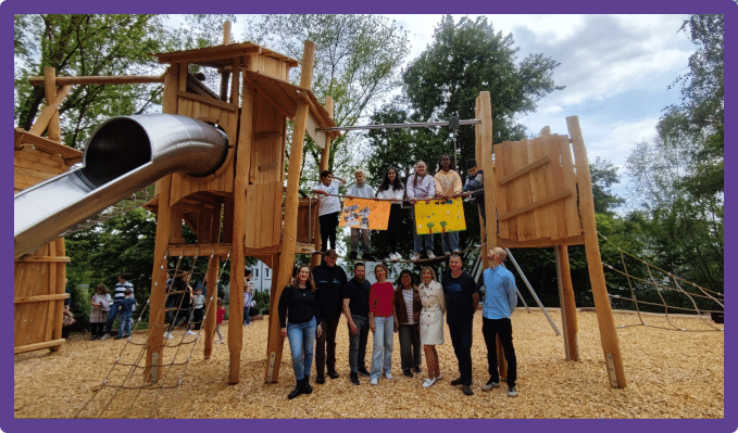 Auf dem Foto ist eine Szene von der Spielplatzeröffnung zu sehen. Eine Kindergruppe, die ihre Ideen eingebracht haben, spielt auf dem neuen Klettergerüst und Erwachsene, die bei der Umsetzung beteiligt waren sind am Fuß des Gerüstes versammelt.