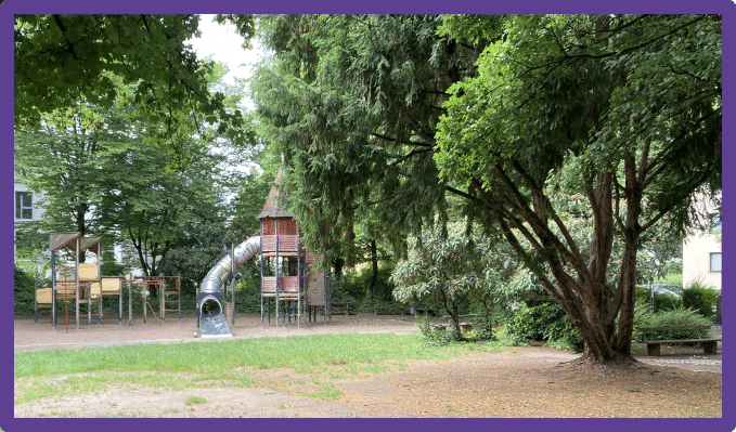 Es ist ein aktuelles Bild vom Spielplatz zu sehen, auf dem sich viele Bäume und Kletterspielgeräte mit Möglichkeit zum Rutschen befinden.